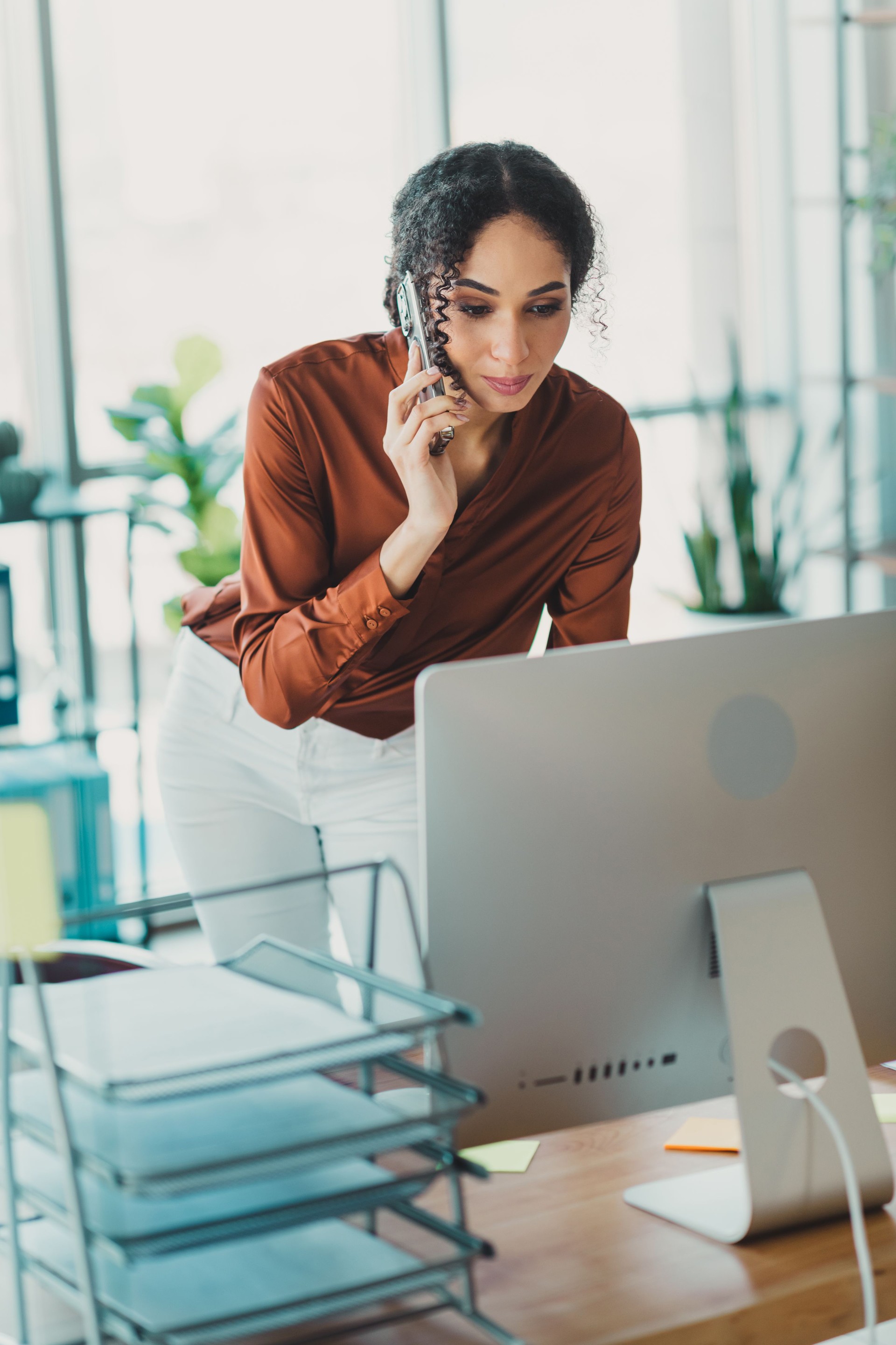 Confident businesswoman engaged in work at her modern office desk, multitasking on a call and computer in a bright, elegant environment